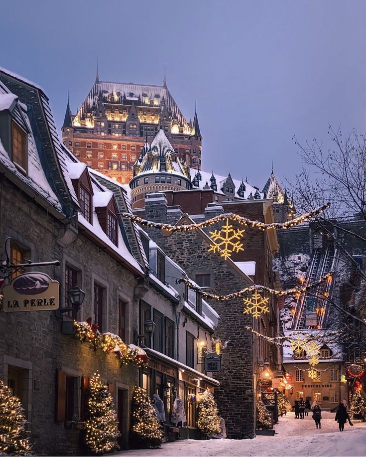 people walk down a snowy street lined with christmas lights and buildings in the background at dusk