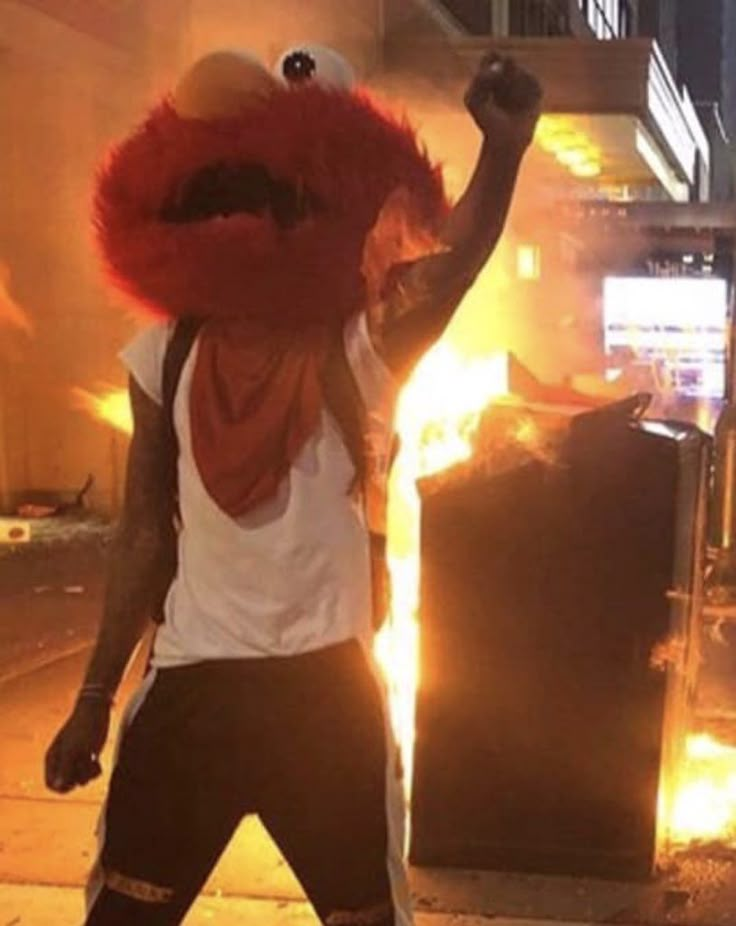 a man wearing a red mask and standing in front of a fire with his arms up