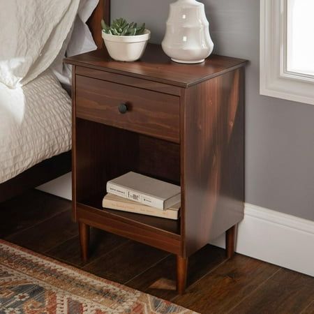 a nightstand with two books on it next to a white vase and a bed in the background