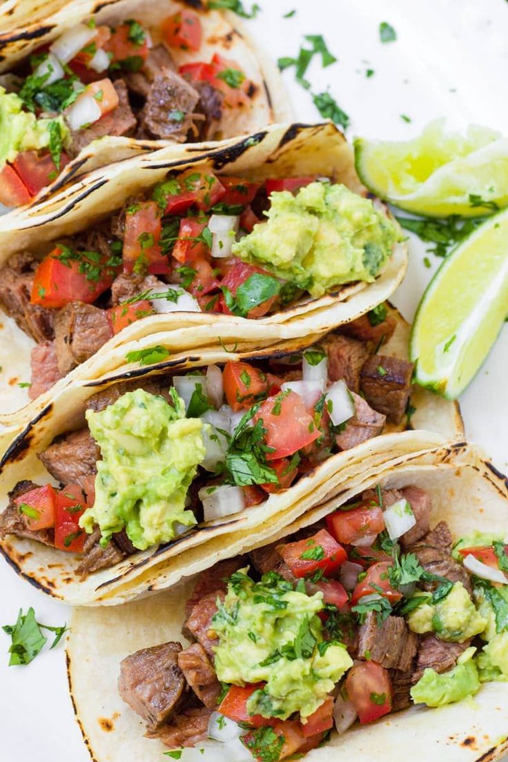 three steak tacos with guacamole, tomatoes and cilantro on the side