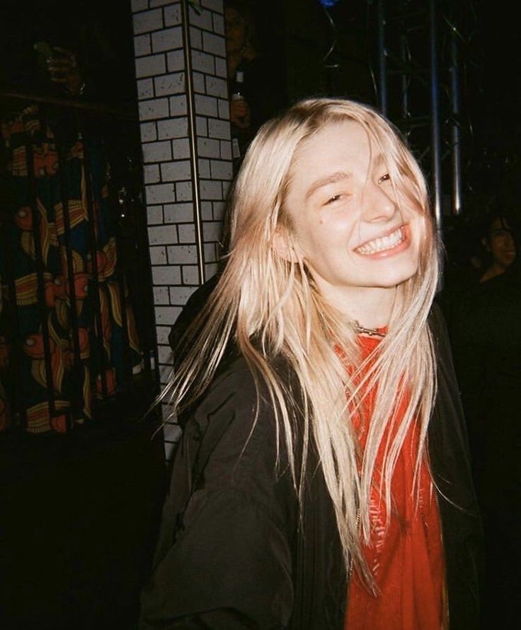 a woman with long blonde hair standing in front of a brick wall and smiling at the camera