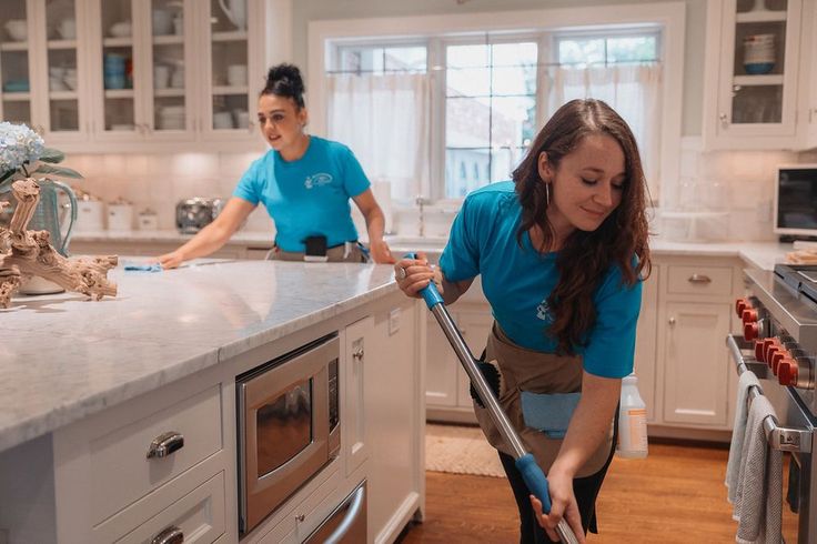 two women in blue shirts are cleaning the kitchen counter top with a vacuum mop