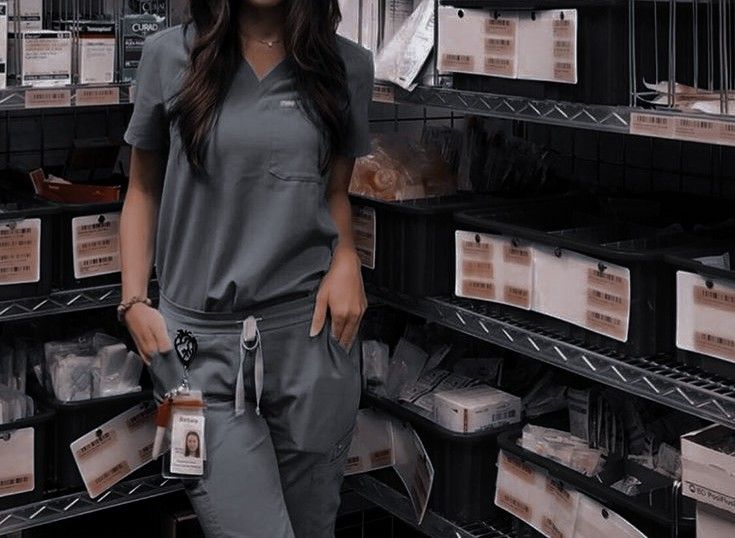 a woman in grey scrubs standing next to shelves with boxes on the wall and she is smiling at the camera