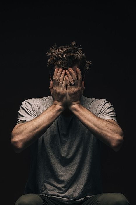 a man sitting on the ground covering his face with both hands, in front of a black background