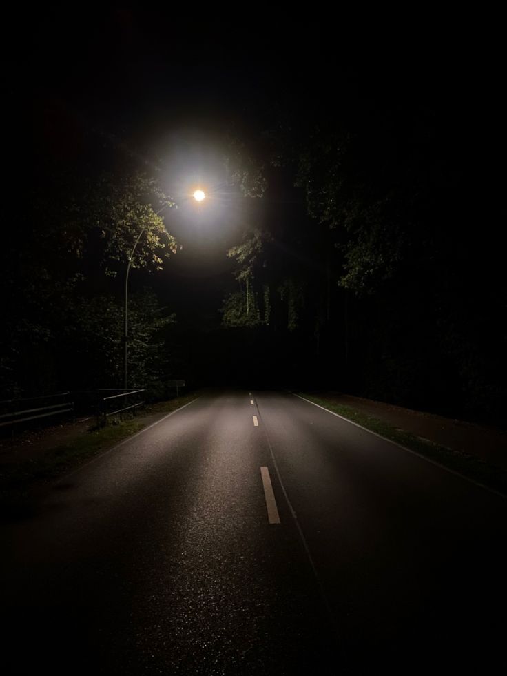 an empty road at night with the light on and street lamp in the dark above