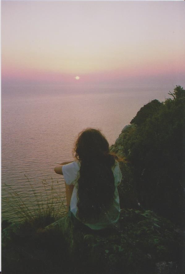 a woman sitting on top of a cliff overlooking the ocean at sunset or dawn with her hair blowing in the wind