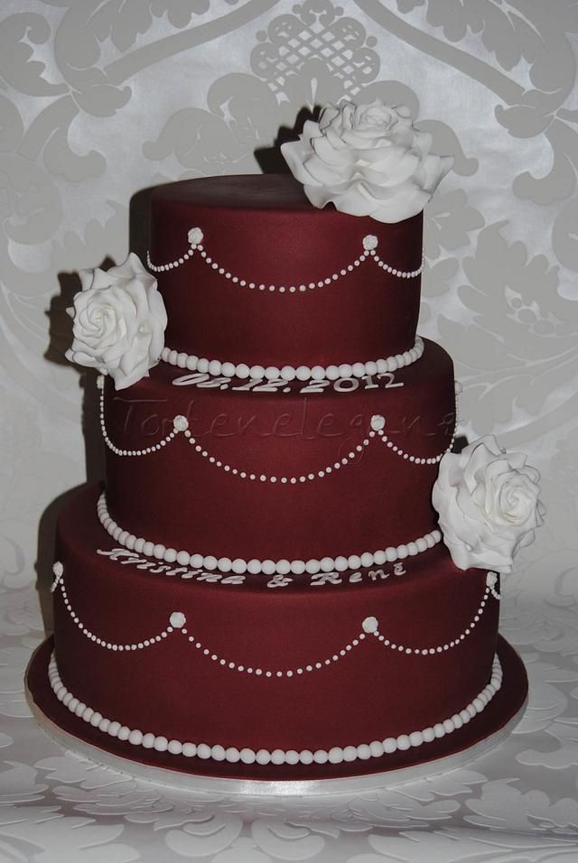 a three tiered red wedding cake with white flowers on the top and pearls around the edges