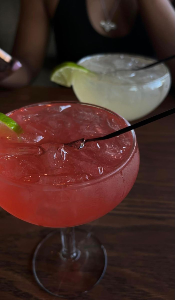 two cocktails sitting on top of a wooden table next to each other, one with a lime wedge in it