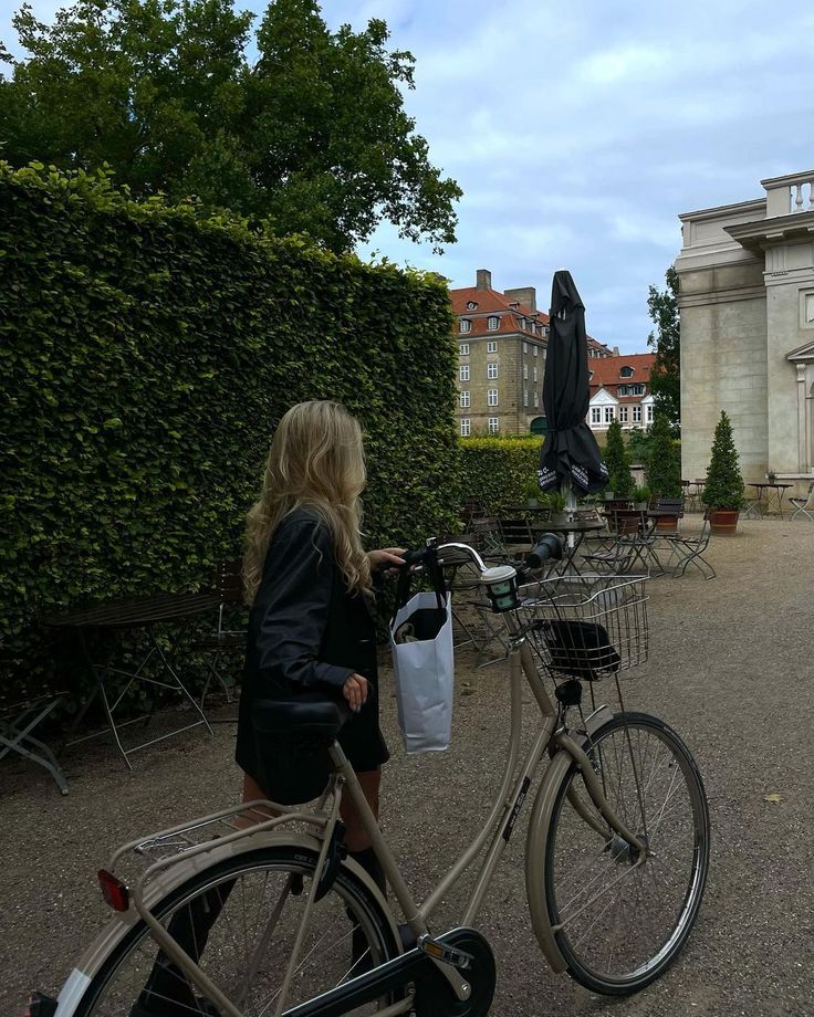 a woman riding a bike down a street next to a tall hedge covered building with an umbrella