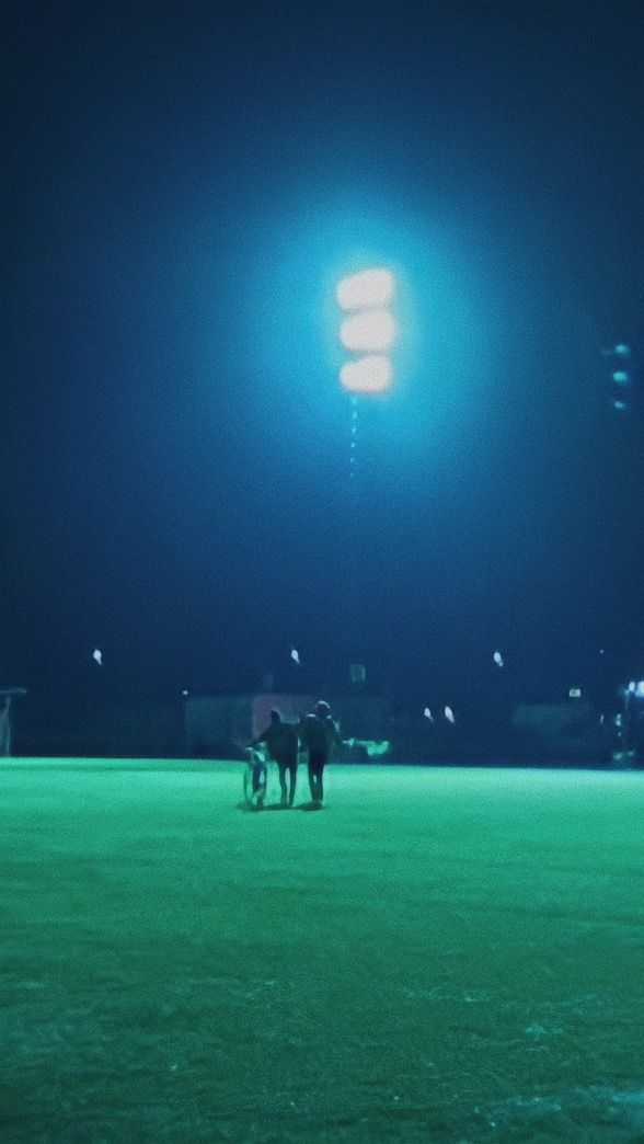 two people standing in the middle of a baseball field at night with lights shining on them