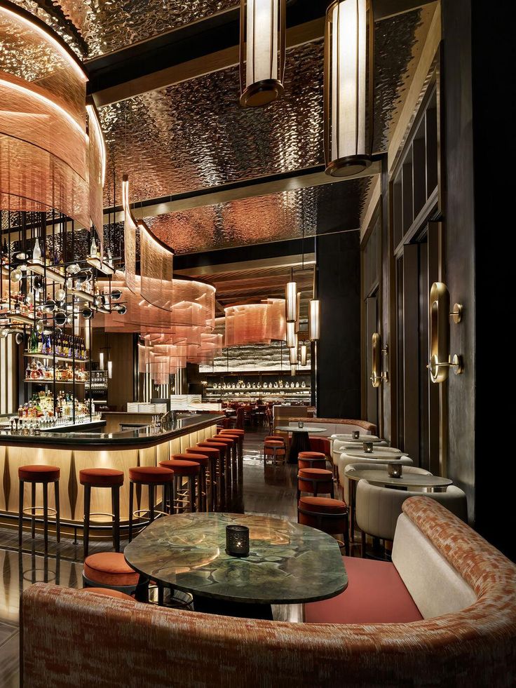 a bar with lots of stools and tables in front of the bar area is lit by chandeliers