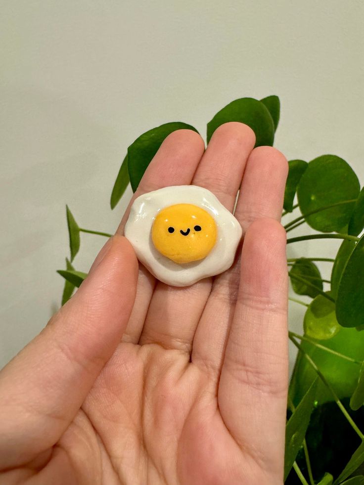 a hand holding a small egg with a smiley face on it's side, next to a potted plant