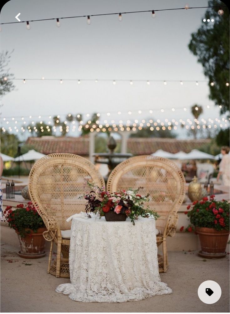 two chairs and a table with flowers on it in front of string lights at an outdoor event