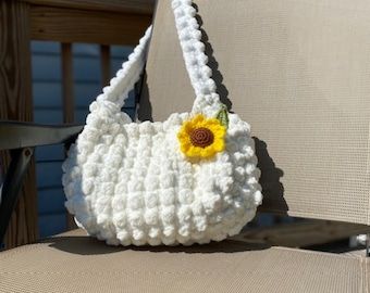 a crocheted white purse with a yellow flower on the front and side, sitting on a chair