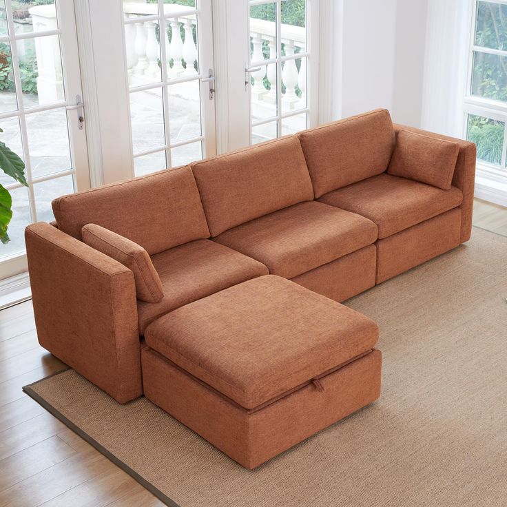 a living room with a large sectional couch and footstool on the floor in front of two windows