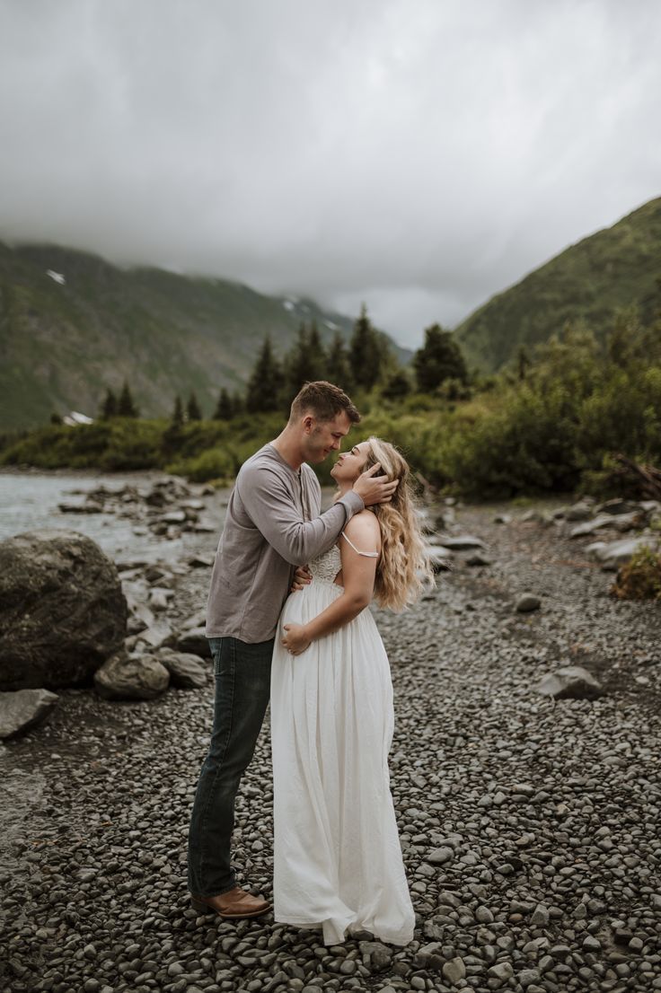 a man and woman standing next to each other on a rocky river bank with mountains in the background