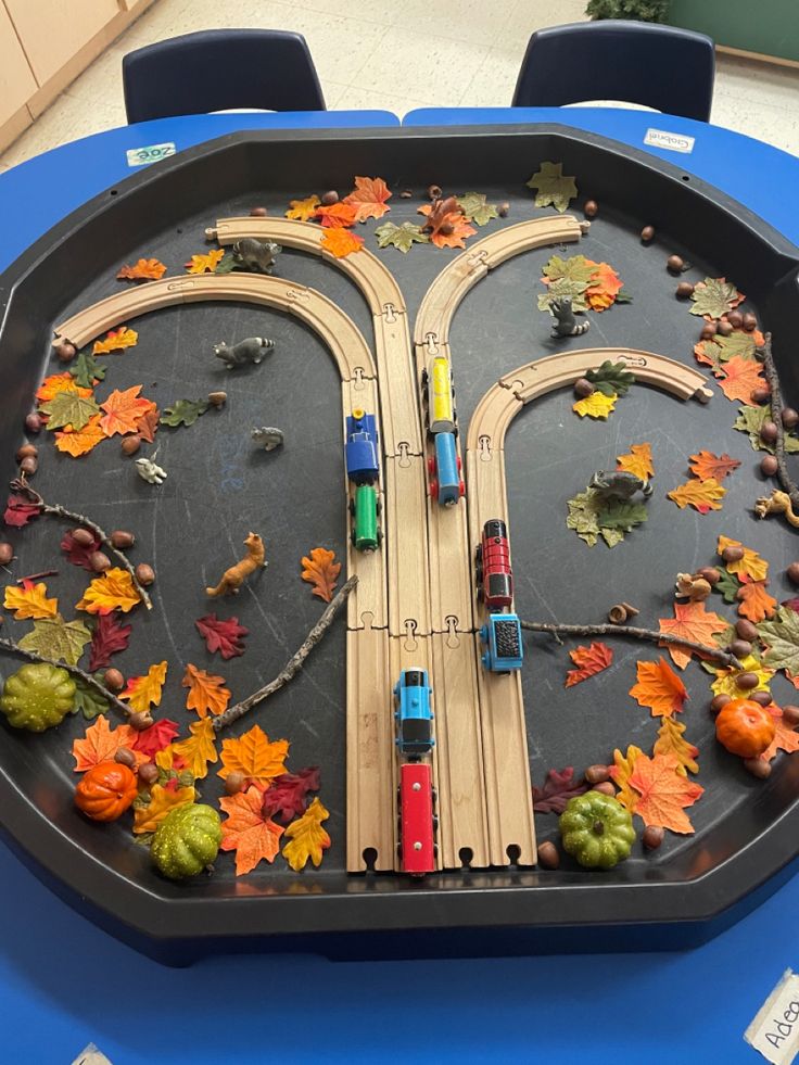 a toy train set on top of a table with fall leaves around it and cars driving down the track