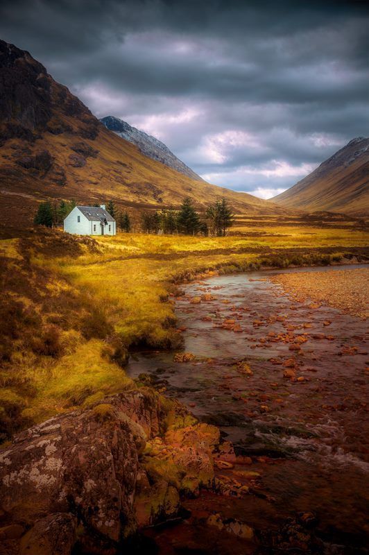 a small house sitting on top of a lush green field next to a river and mountains