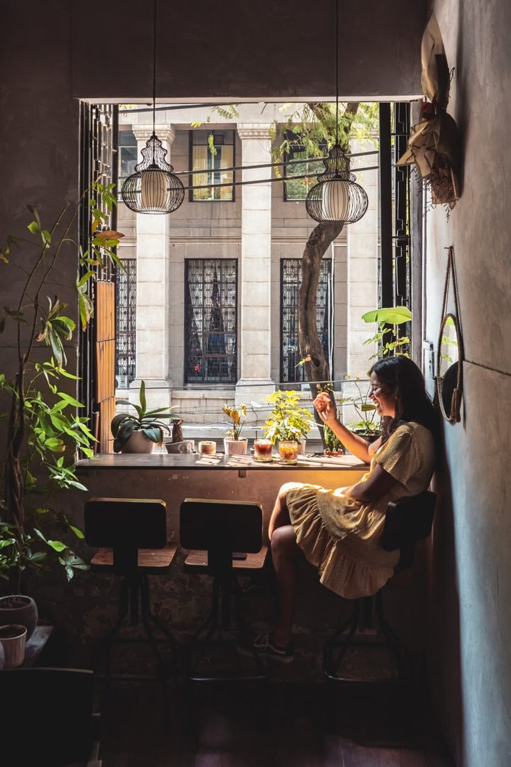 a woman sitting at a table in front of a window with potted plants on it