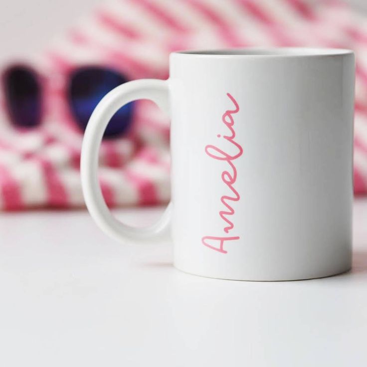 a white coffee mug with pink lettering sitting on a table next to a red and white striped pillow