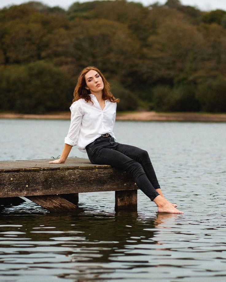 a woman sitting on top of a wooden dock next to the water with trees in the background