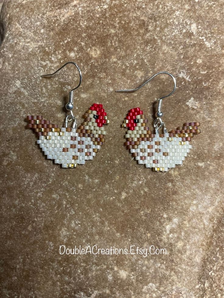 a pair of chicken earrings on top of a stone slab with red and white beads