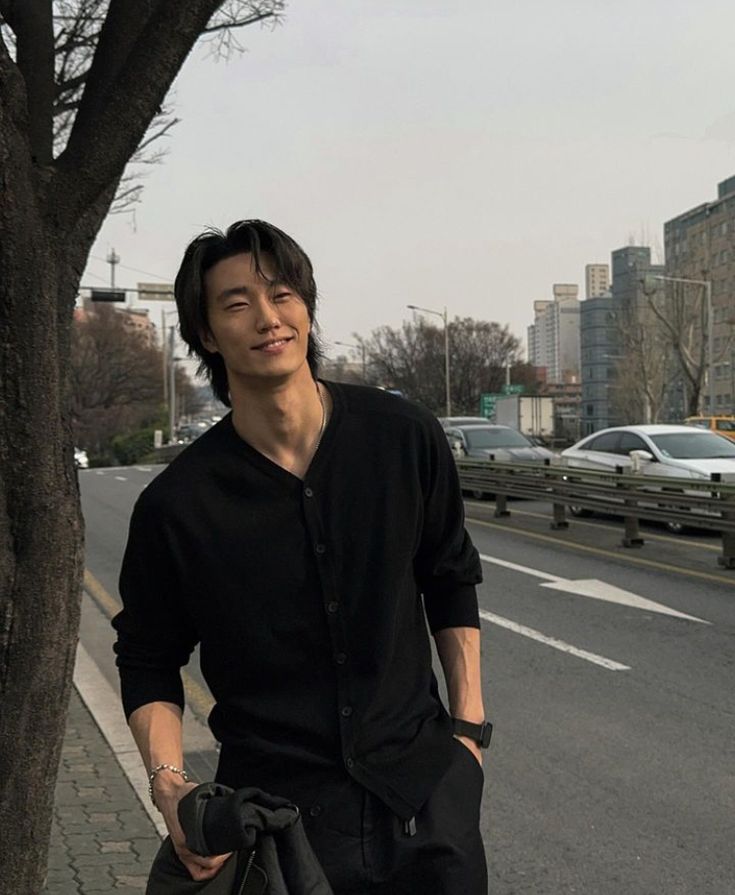 a man standing next to a tree on the side of a road with his hands in his pockets