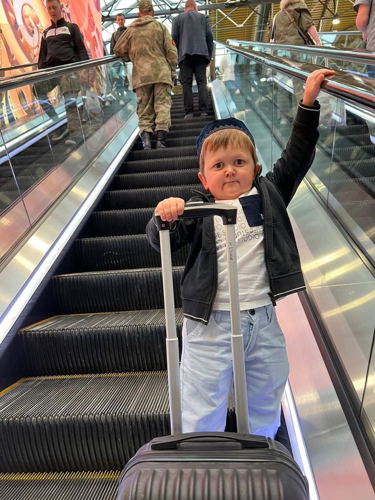 a little boy is standing on an escalator with his luggage and waving at the camera