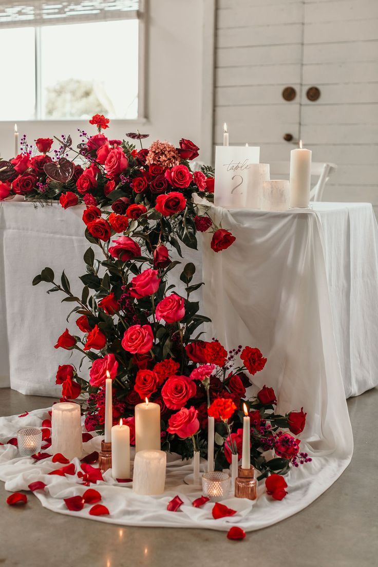 red roses and candles are arranged on a table with white linens in the background