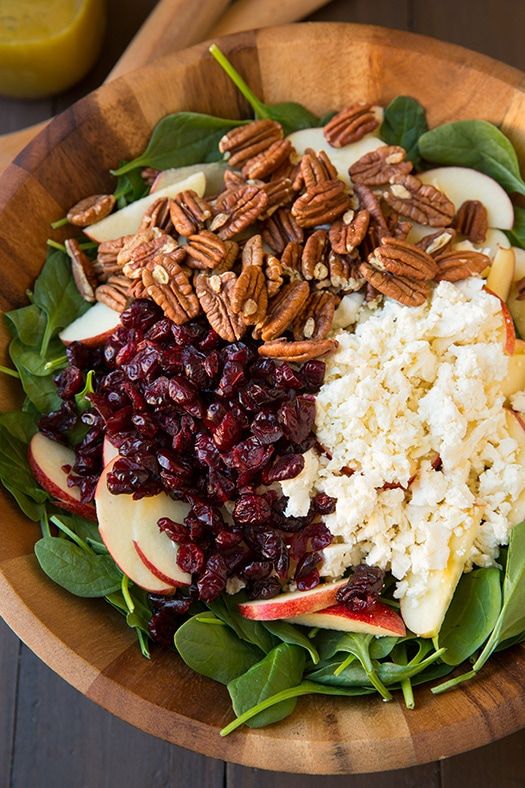 a wooden bowl filled with apples, cranberries and pecans on top of spinach