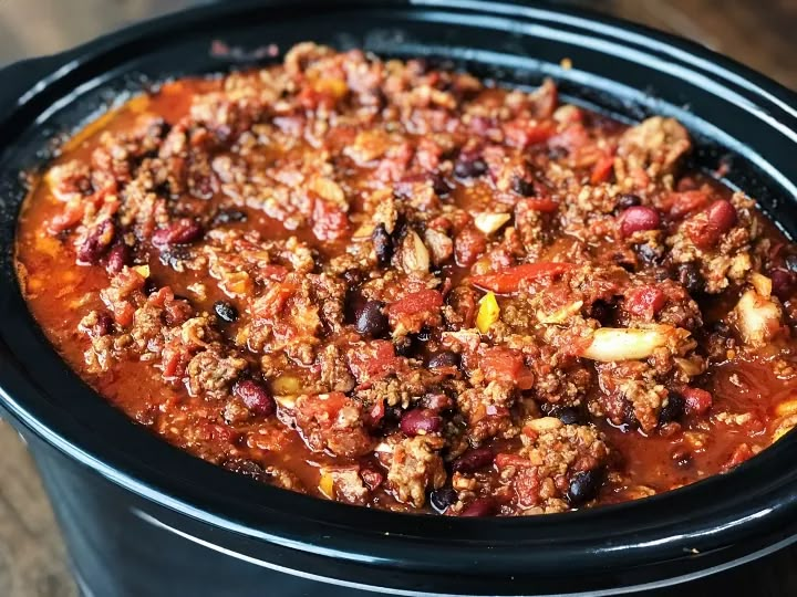 a crock pot filled with chili and meat in it sitting on a wooden table