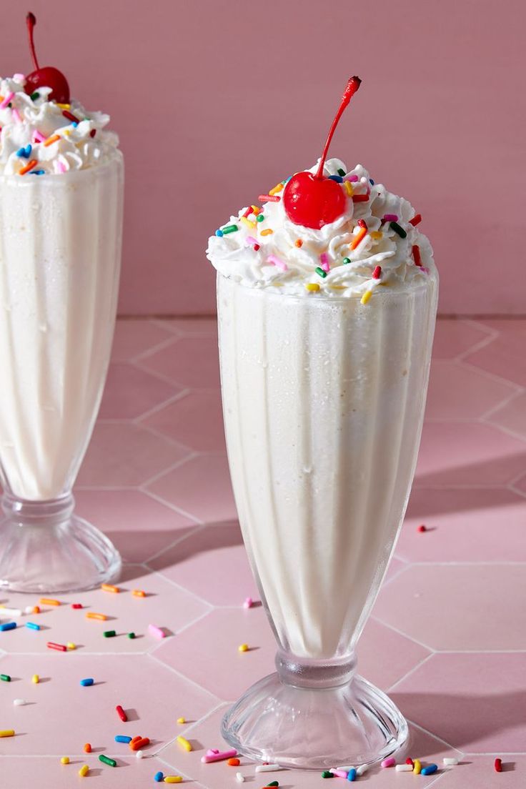 two milkshakes with whipped cream and sprinkles on pink tablecloth