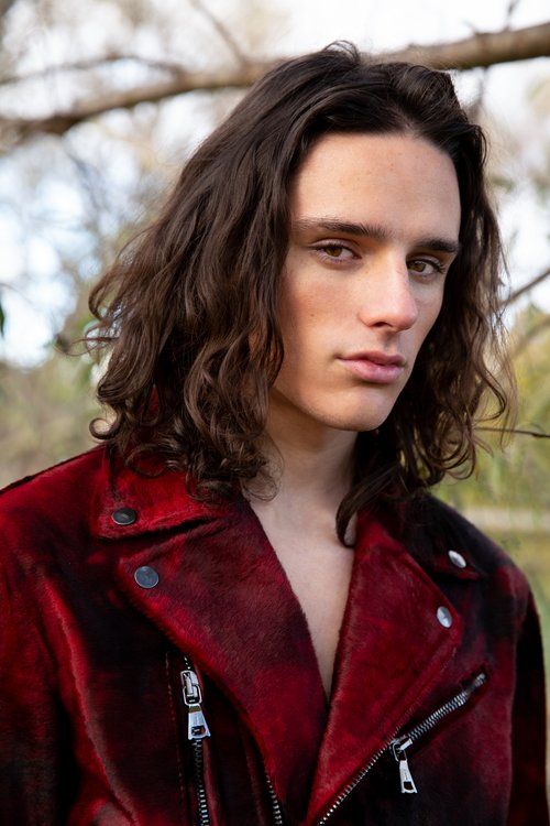 a man with long hair wearing a red jacket and looking at the camera while standing in front of a tree