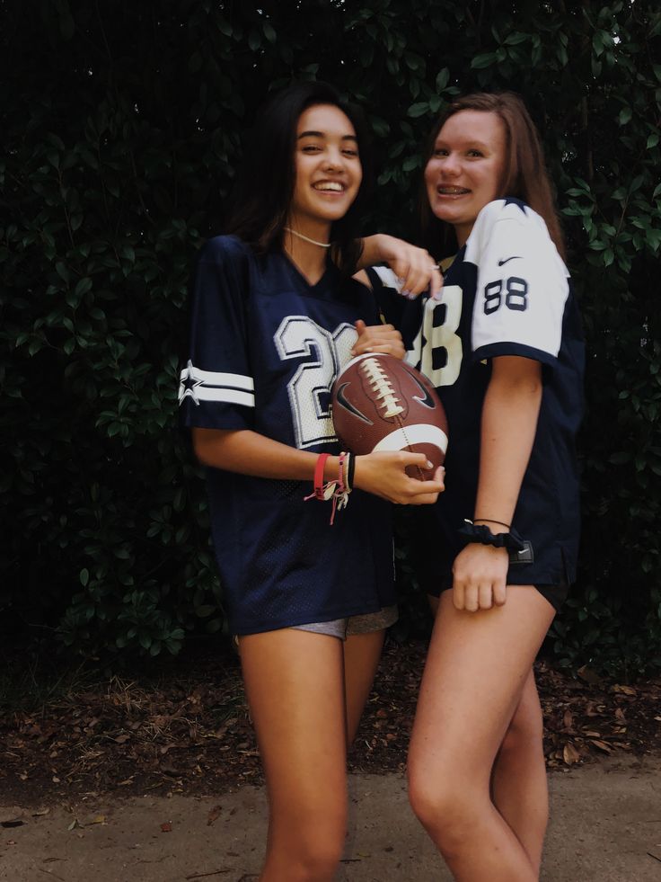 two girls in football uniforms posing for the camera with one holding a football and the other wearing shorts