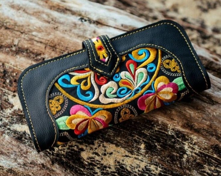 an embroidered wallet sitting on top of a piece of wooden plank with colorful flowers and leaves