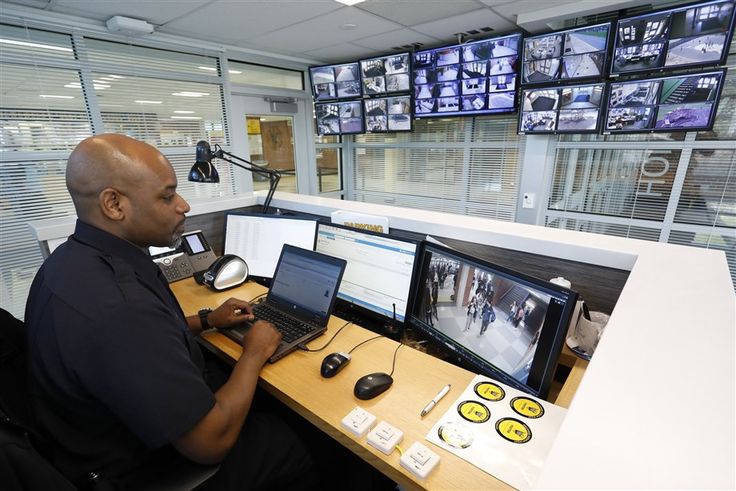 a man sitting at a desk in front of two computer monitors with multiple screens on the wall behind him