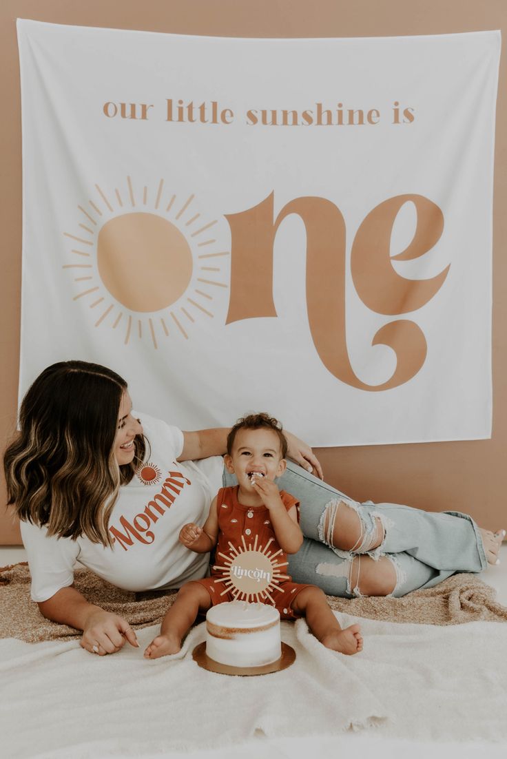 two women and a baby sitting in front of a one year birthday banner with the words our little sunshine is one on it