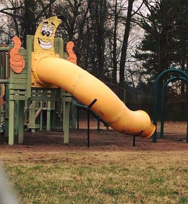 a yellow slide in the middle of a park with trees behind it and a banana on top