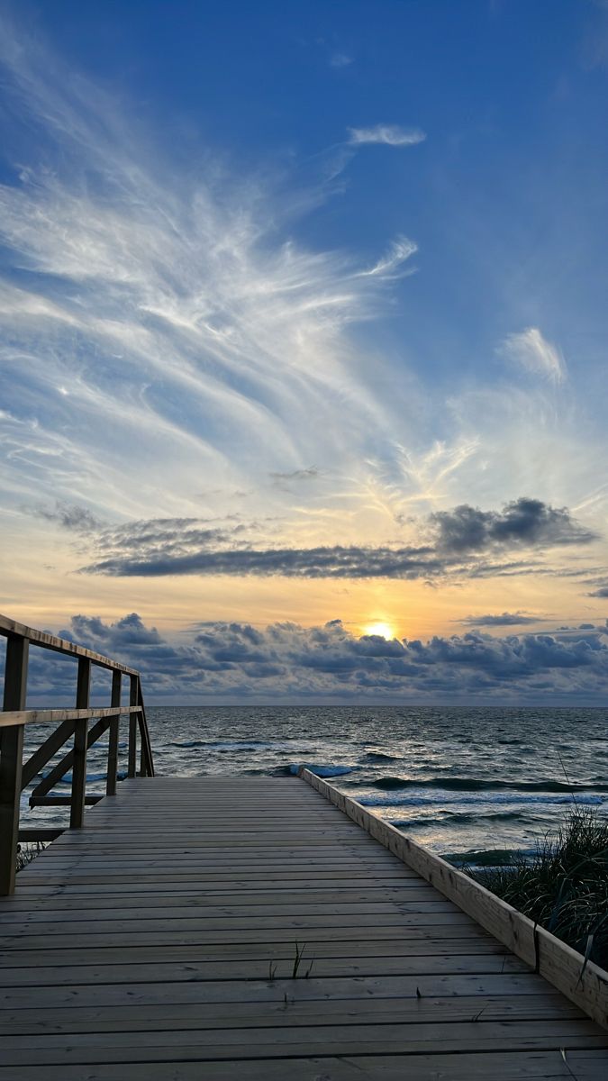 a wooden walkway leading to the ocean with clouds in the sky and sun going down