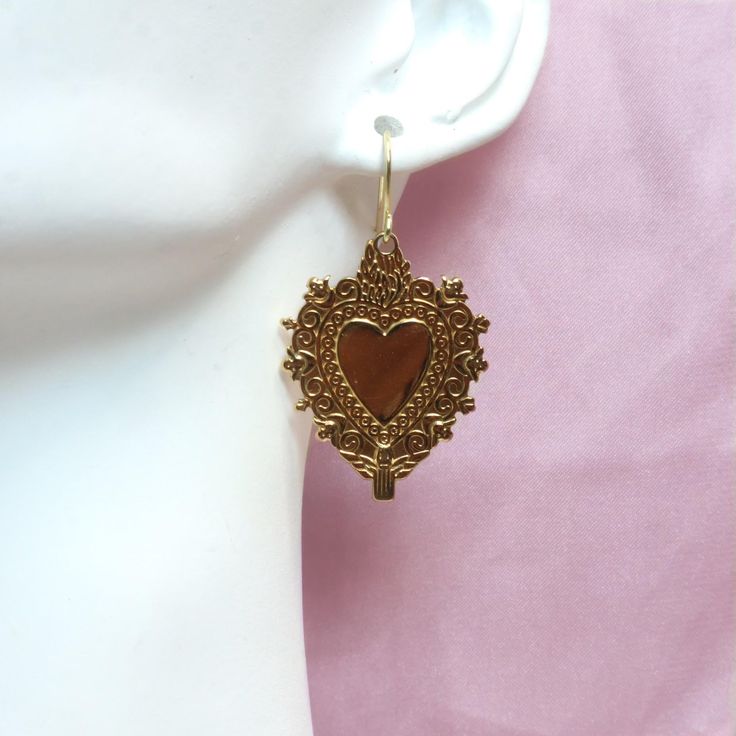 The Divine Heart Earrings feature gold-colored stainless steel pendants inspired by the iconic Sacred Heart imagery. The hooks are crafted from 14k hypoallergenic gold, while the pendants are made from tarnish-resistant stainless steel, ensuring both comfort and durability. Perfect for those who appreciate meaningful design and lasting quality. 14k Hypoallergenic Gold Hooks Earring Pendant Measurement: 30x23mm Tarnish Resistant With Love From Texas Nickel Free Brass Heart-shaped Jewelry, Nickel Free Heart-shaped Brass Jewelry, Nickel Free Heart Shaped Brass Jewelry, Nickel-free Heart-shaped Brass Jewelry, Gold Heart-shaped Plug Earrings As Gift, Heart-shaped Gold Plug Earrings As Gift, Gold Heart Shaped Plug Earrings As Gift, Heart Shaped Gold Plug Earrings As Gift, Gold Teardrop Heart Charm Earrings