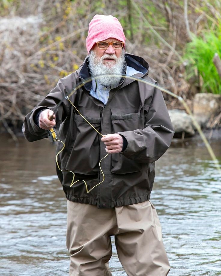 an old man standing in the water holding a fishing rod and wearing a pink hat