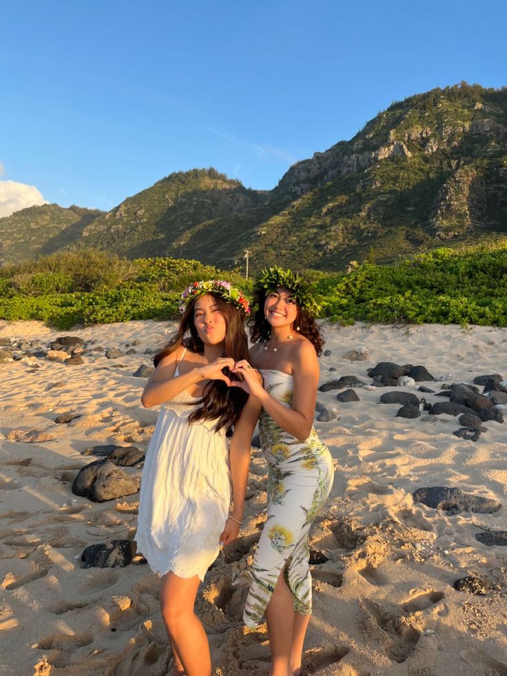 two young women standing on top of a sandy beach next to the ocean with mountains in the background