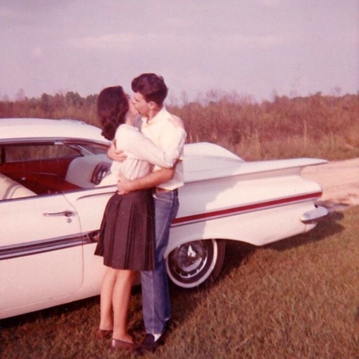 a man and woman standing next to an old car in the grass with their arms around each other