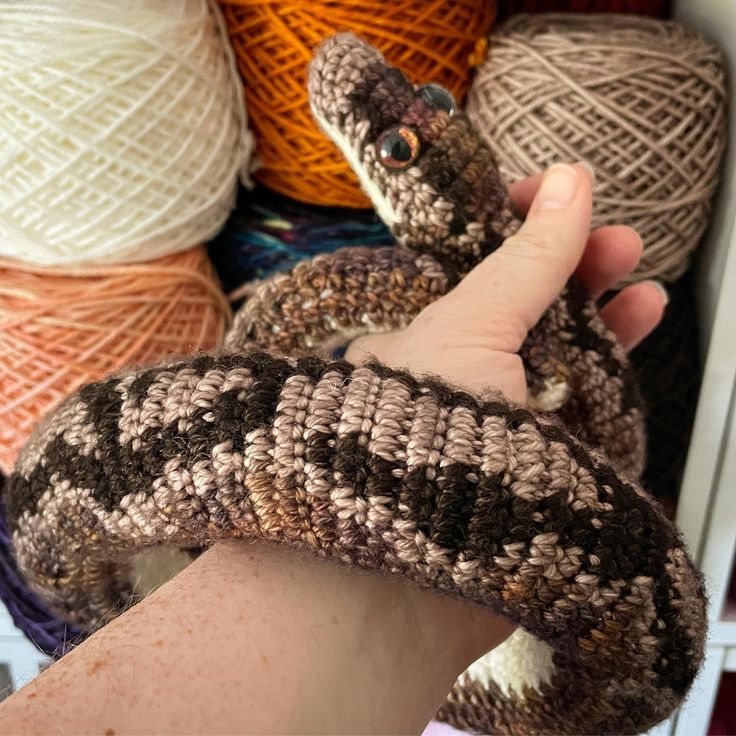 a hand is holding a knitted snake in front of balls of yarn