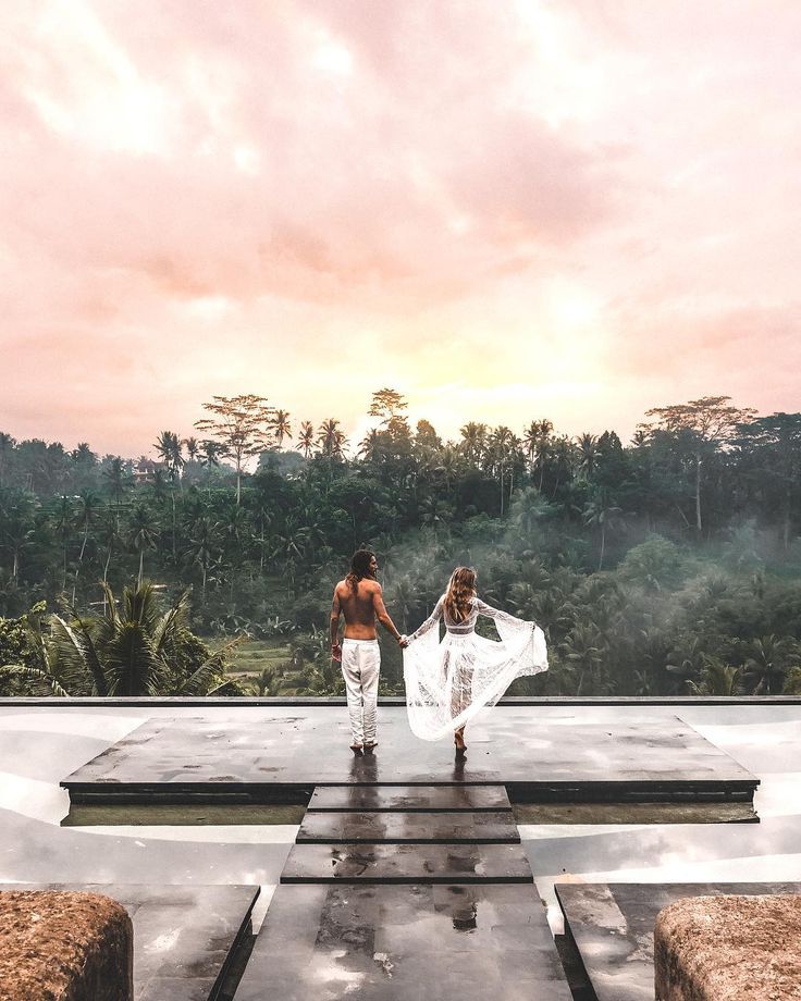 two women in white dresses walking down a walkway with trees and clouds behind them at sunset