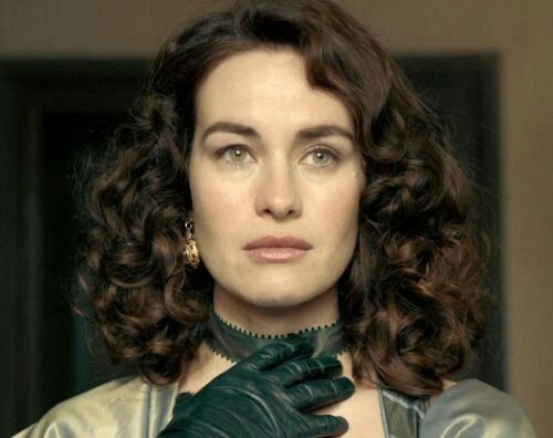 a woman with long curly hair wearing black gloves and a green dress is looking at the camera