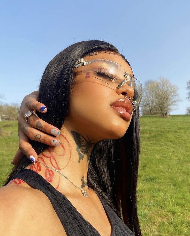 a woman with tattoos on her neck and glasses is posing for the camera in front of a grassy field