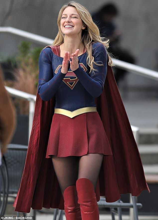 a woman in a skirt and cape smiles as she stands on the set of supergirl