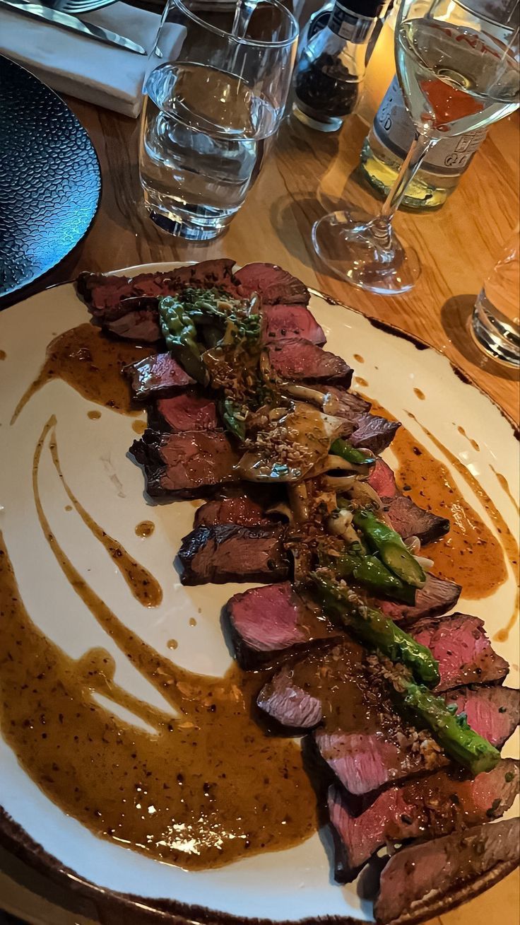 a white plate topped with meat and vegetables on top of a wooden table next to wine glasses