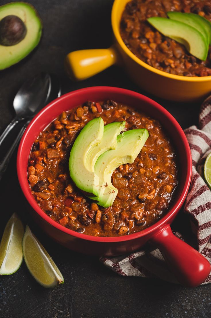 two bowls filled with chili and avocado on top of a table next to spoons